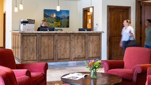 Een receptie met een houten balie, een receptioniste en een groot landschap op de muur. Op de voorgrond staan rode fauteuils en een bijzettafeltje met een bloevas.