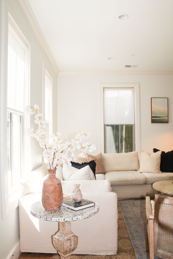 A living room with a white sofa, a round table with a vase and book, and a large window.