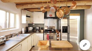 A kitchen with wooden ceiling beams, hanging pots, stainless steel appliances, and a wooden cutting board on the counter.