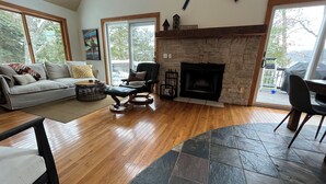 A spacious living room with a stone fireplace, wooden flooring, and large windows offering a view of the outdoors. - Beautiful Cottage on Private Lake close to Manistee  (Wellston)