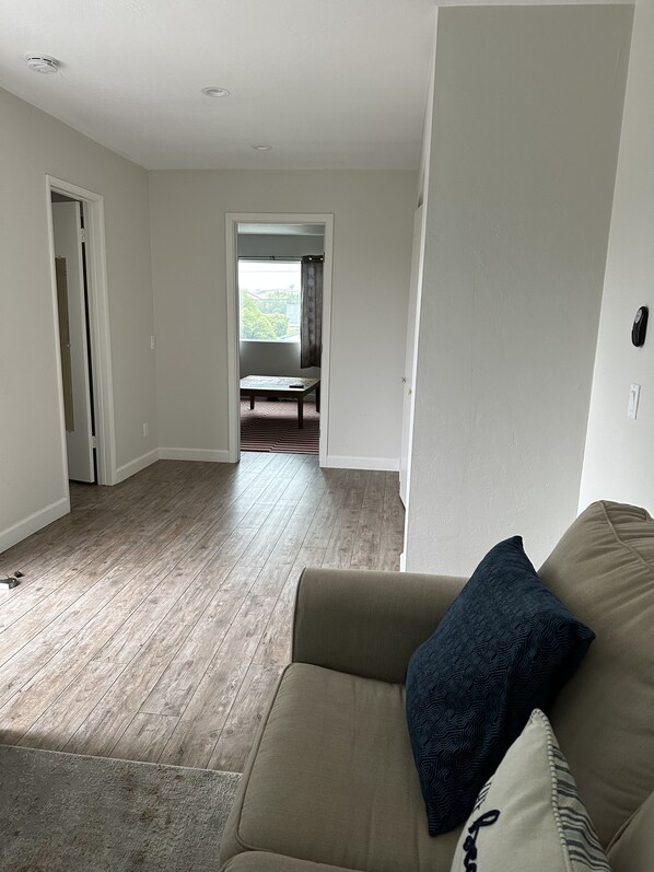 A living room with a beige sofa, a patterned throw pillow, and a wooden floor. There is a doorway leading to a balcony with a view of greenery. - Carlsbad Getaway - Spacious and new! (Carlsbad)