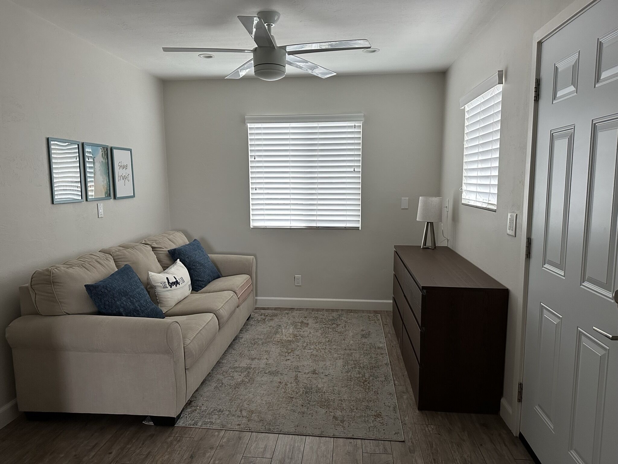 Ein neutral eingerichtetes Wohnzimmer mit beiger Couch, Deckenventilator, Fenster mit Jalousien und einem Schminktisch.