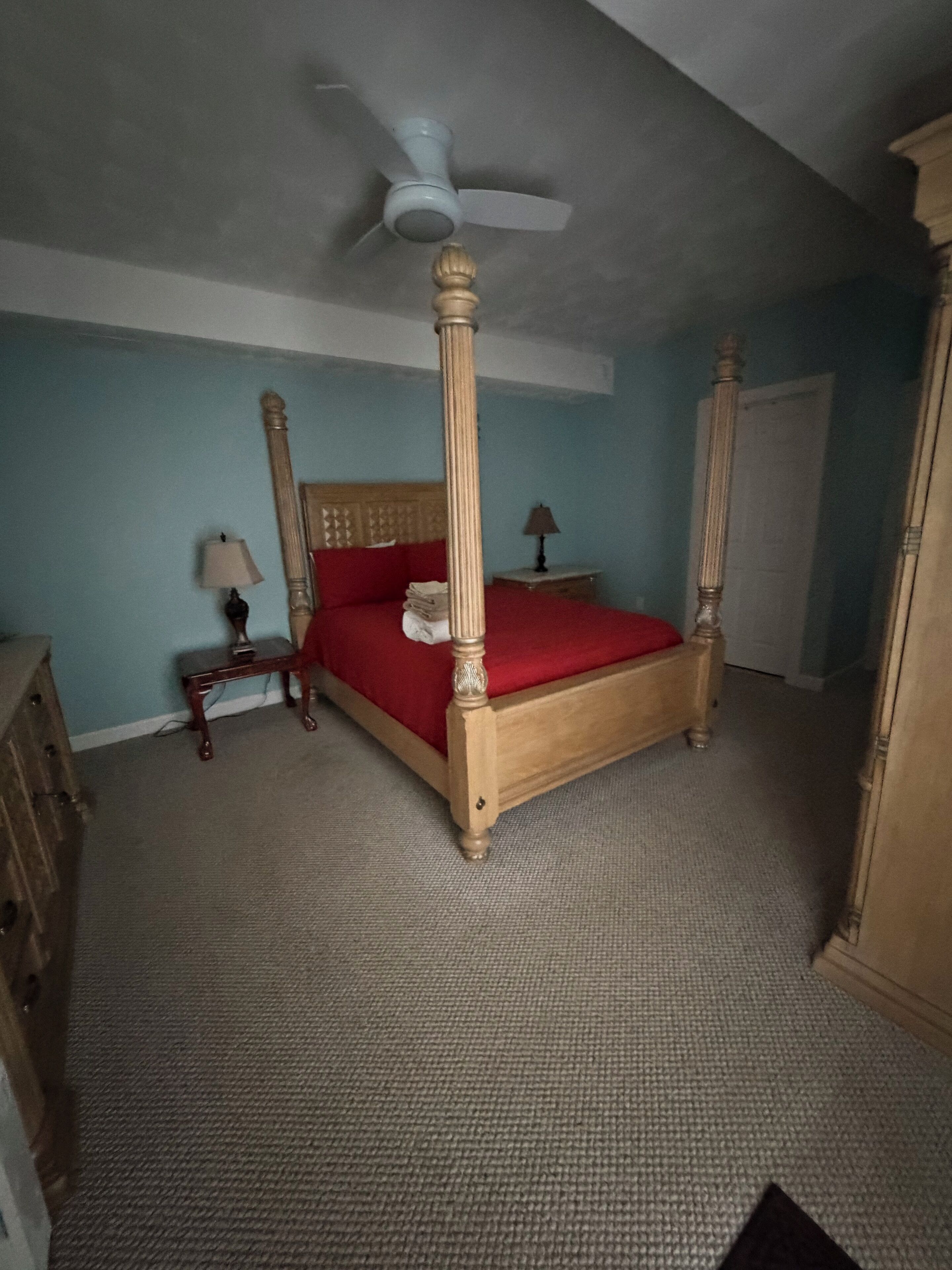 A four-poster bed with a wooden frame and red bedding, a ceiling fan, a nightstand with a lamp, and a dresser.