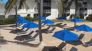 A beachside hotel with multiple balconies, palm trees, and blue umbrellas. - Simply the Best at the Most Reasonable Price-Cayman Reef Resort #2 (George Town)