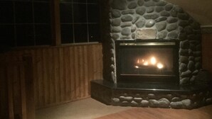 A stone fireplace with a fire, wooden walls, and a window with a grid pattern. - Large cozy modern cabin  (Centennial)