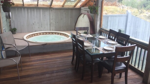 A covered patio with a hot tub, dining table, chairs, and a view of a rural landscape. - Large cozy modern cabin  (Centennial)