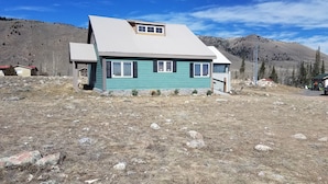 A single-story house with a gabled roof and a small porch, surrounded by a dry, rocky landscape with mountains in the background. - Large cozy modern cabin  (Centennial)