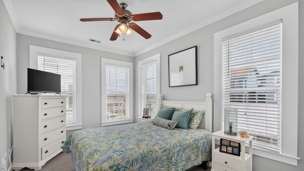 A bedroom with a bed, dresser, TV, ceiling fan, and windows with blinds.