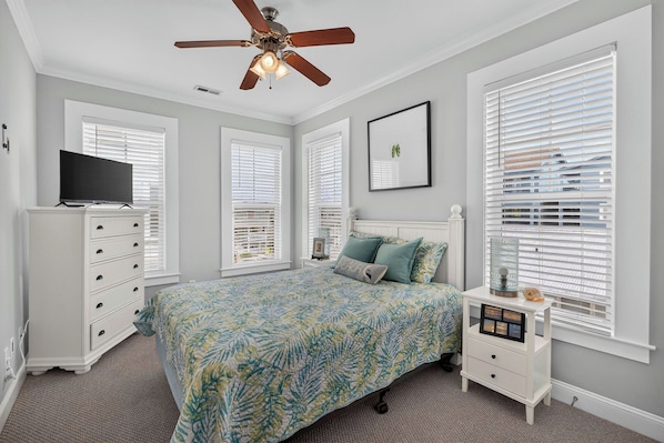 Une chambre à coucher comprenant un lit, une commode, un téléviseur, un ventilateur de plafond et des fenêtres équipées de stores.