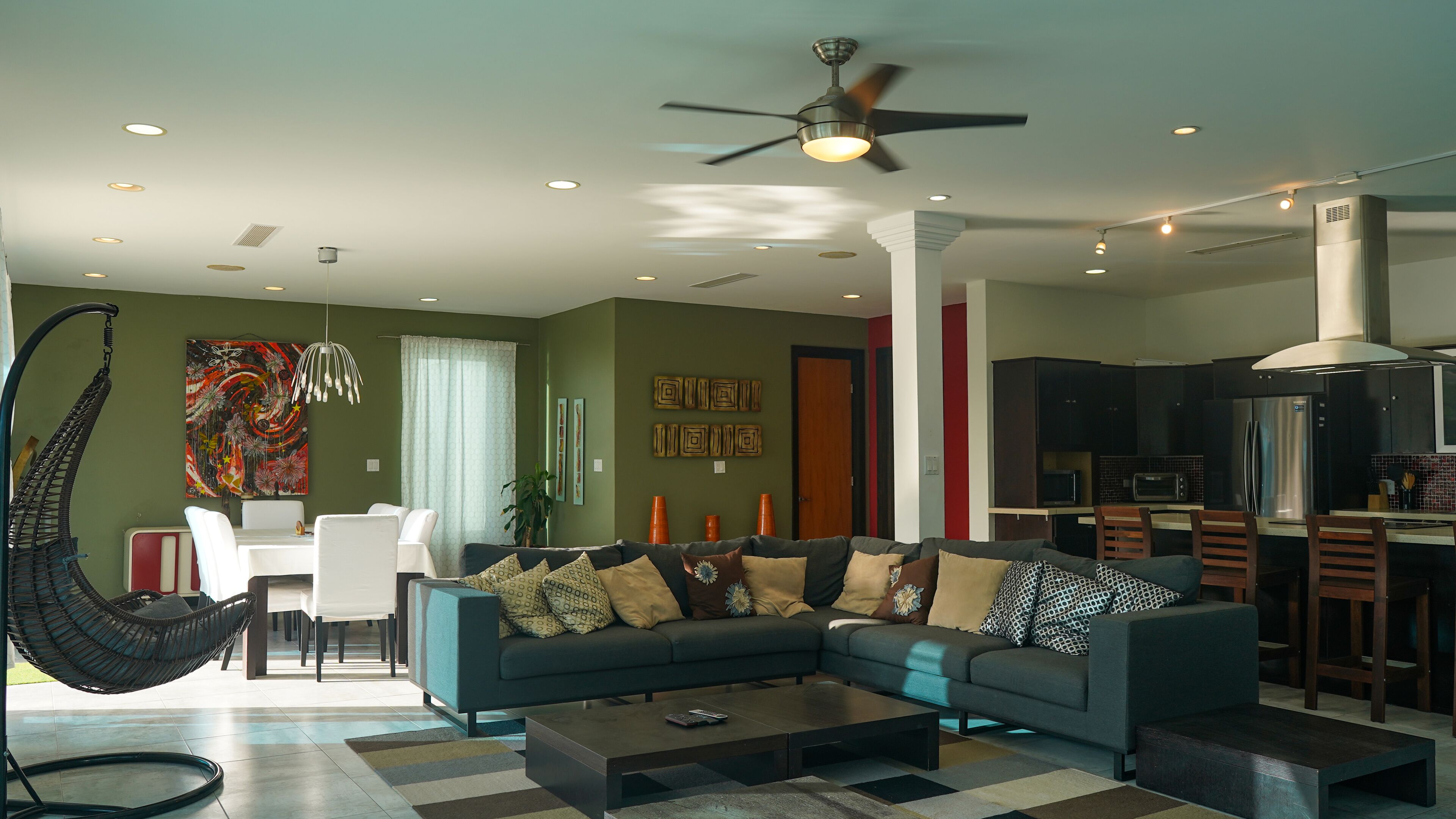 A modern living room with a sectional sofa, a hanging chair, a dining area, and a kitchen with dark cabinetry.