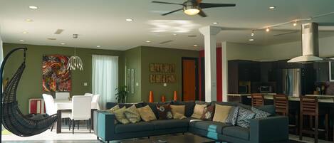 A modern living room with a sectional sofa, a hanging chair, a dining area, and a kitchen with dark cabinetry.