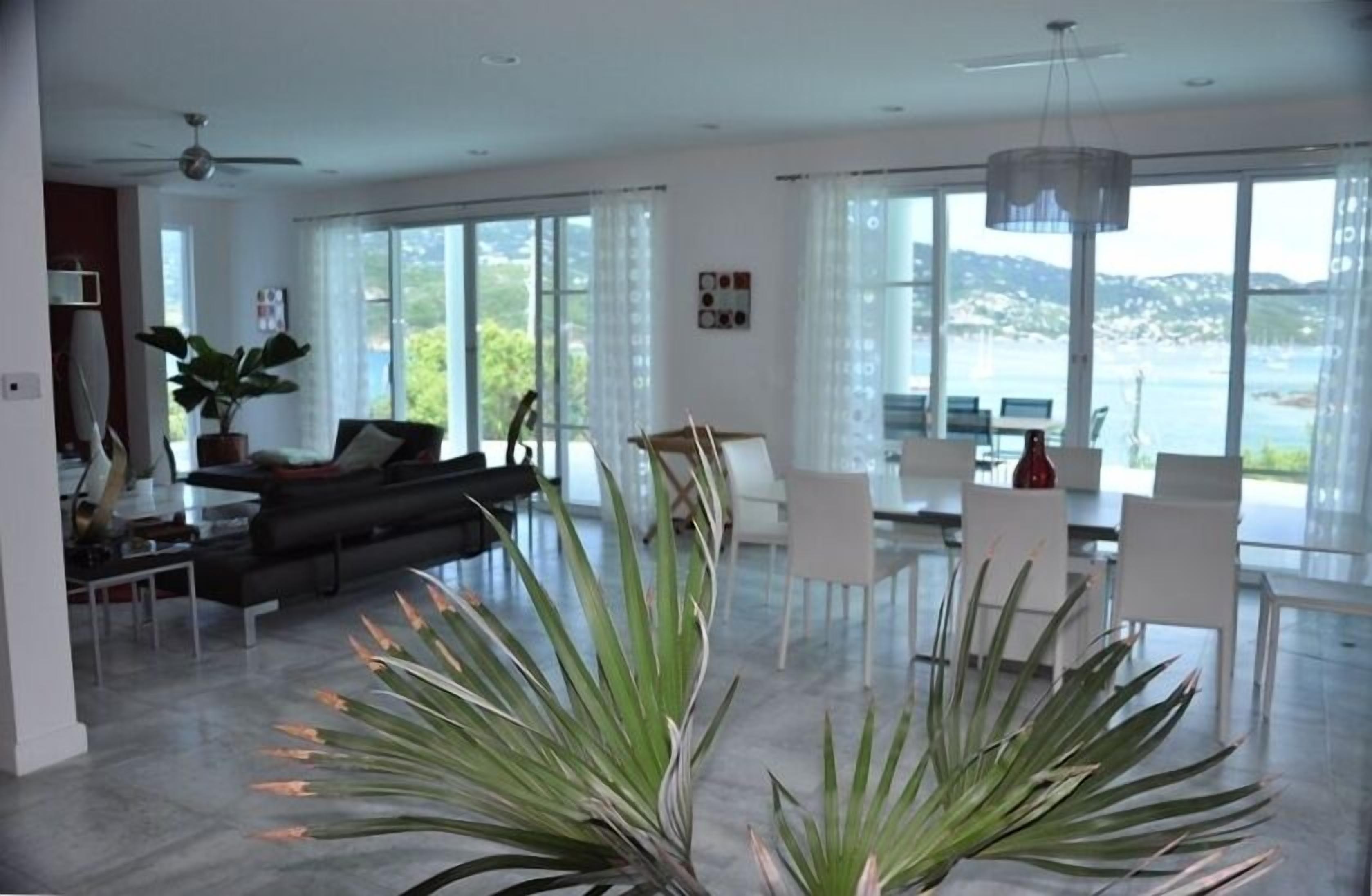 A spacious living room with a large window, a ceiling fan, a dining area with white chairs, and a black sofa.