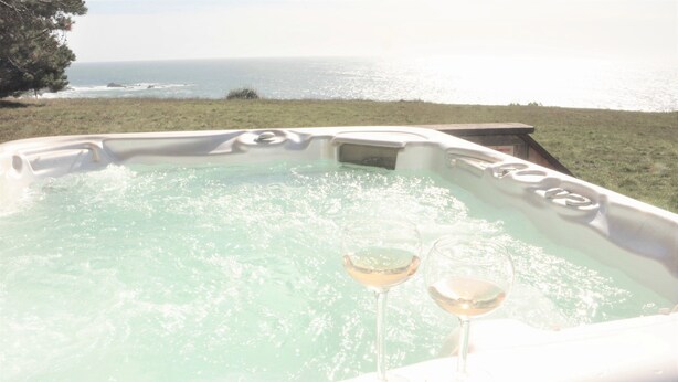 A hot tub with two glasses of white wine, overlooking a grassy area and the ocean.