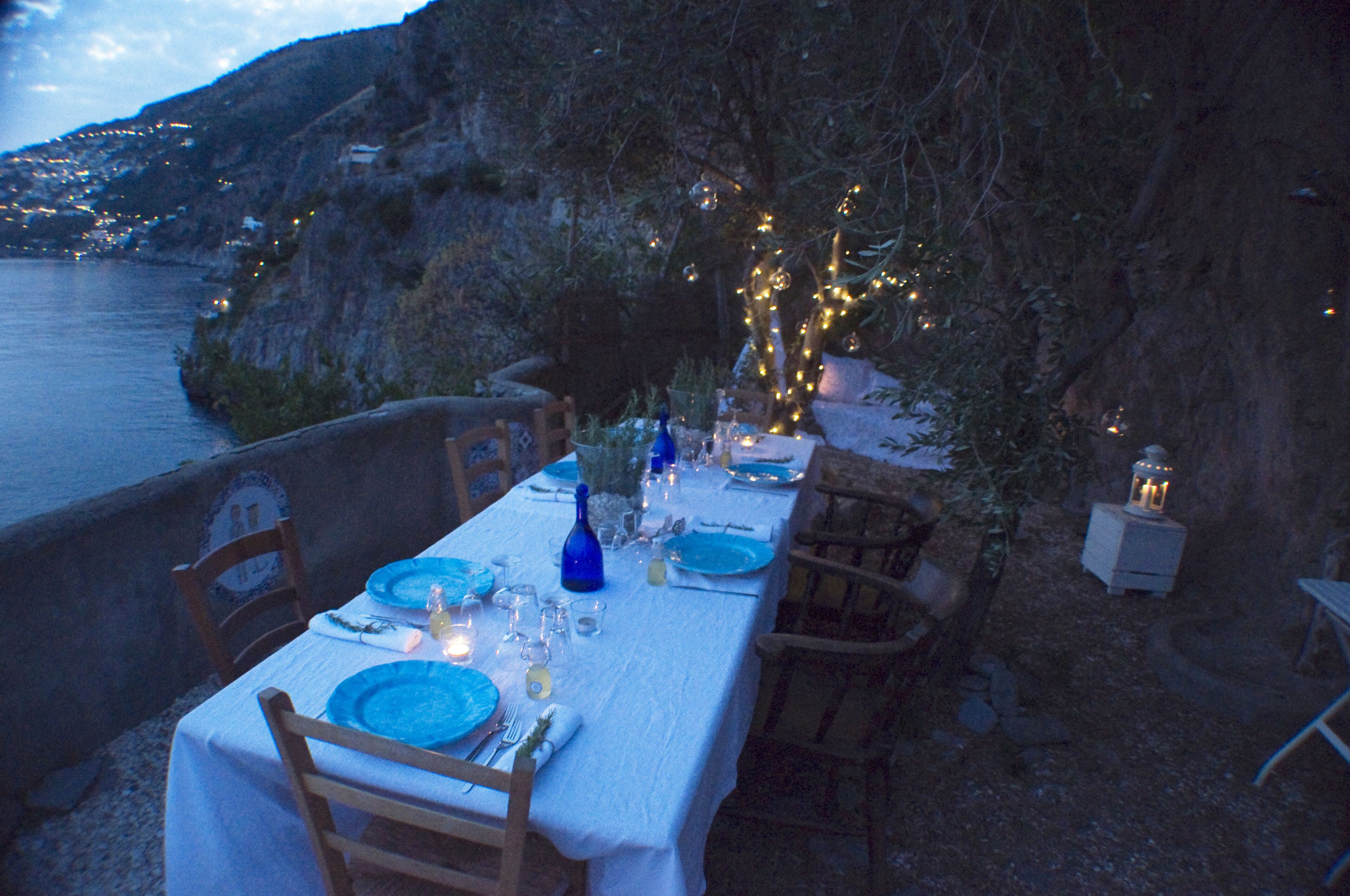 Ein Tisch im Freien mit blauem Geschirr, Gläsern und Kerzen, mit Blick auf eine Küstenlandschaft und einem Berg im Hintergrund.