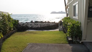 Une vue sur la côte, avec une zone herbeuse qui mène à un rivage rocheux, un bâtiment doté d’un balcon donnant sur la mer, et un navire lointain à l’horizon.
