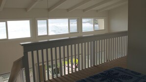 Une chambre avec vue sur l’océan, grâce à de grandes fenêtres, un balcon doté d’une balustrade blanche et une couvre-lit à motifs.