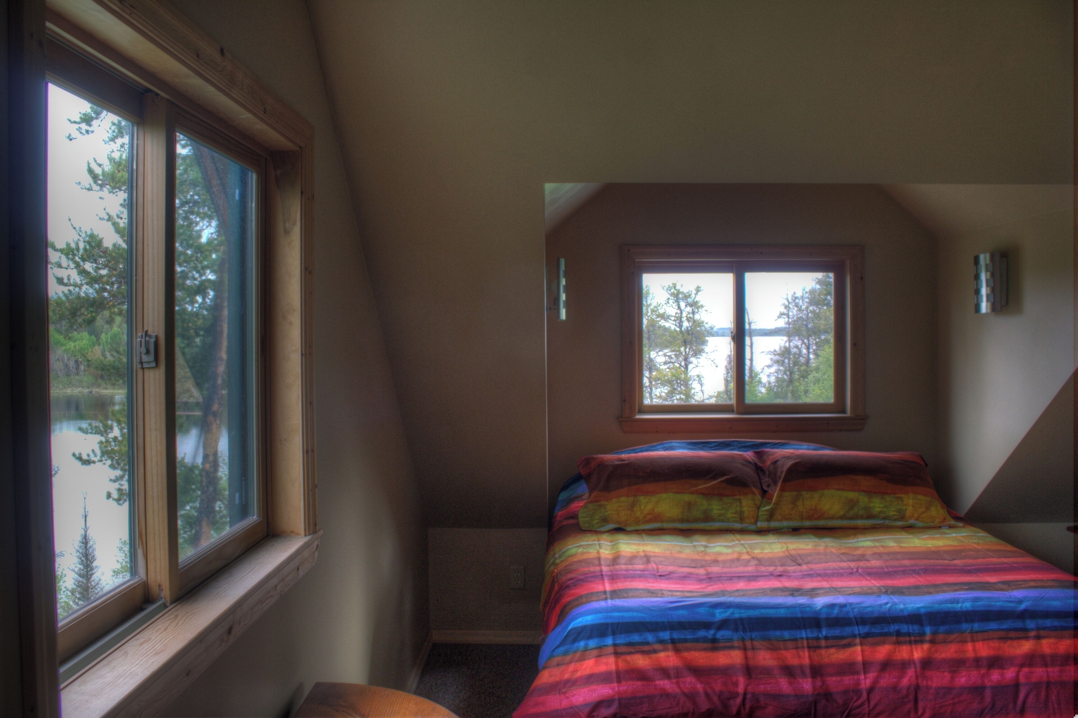 Een slaapkamer met een groot raam, een bed met een kleurrijke gestreepte dekbedovertrek en een klein nachtkastje.