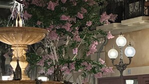 Ein Brunnen, aus dessen Mitte Wasser sprudelt, umgeben von blühenden, rosa Pflanzen.