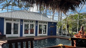 Zona de piscina con terraza de madera, techo de paja y un edificio con puertas de vidrio y fachada azul.
