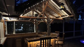 Zona de bar al aire libre con techo de paja, muebles de madera y vistas a la ciudad de noche.