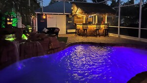 Un área de piscina con bar de techo de paja y sillas, rodeada de palmeras y un espacio de estar cubierto.