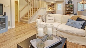 Un salon moderne avec un escalier, un canapé blanc agrémenté de coussins, une table basse avec des bougies et une bibliothèque.