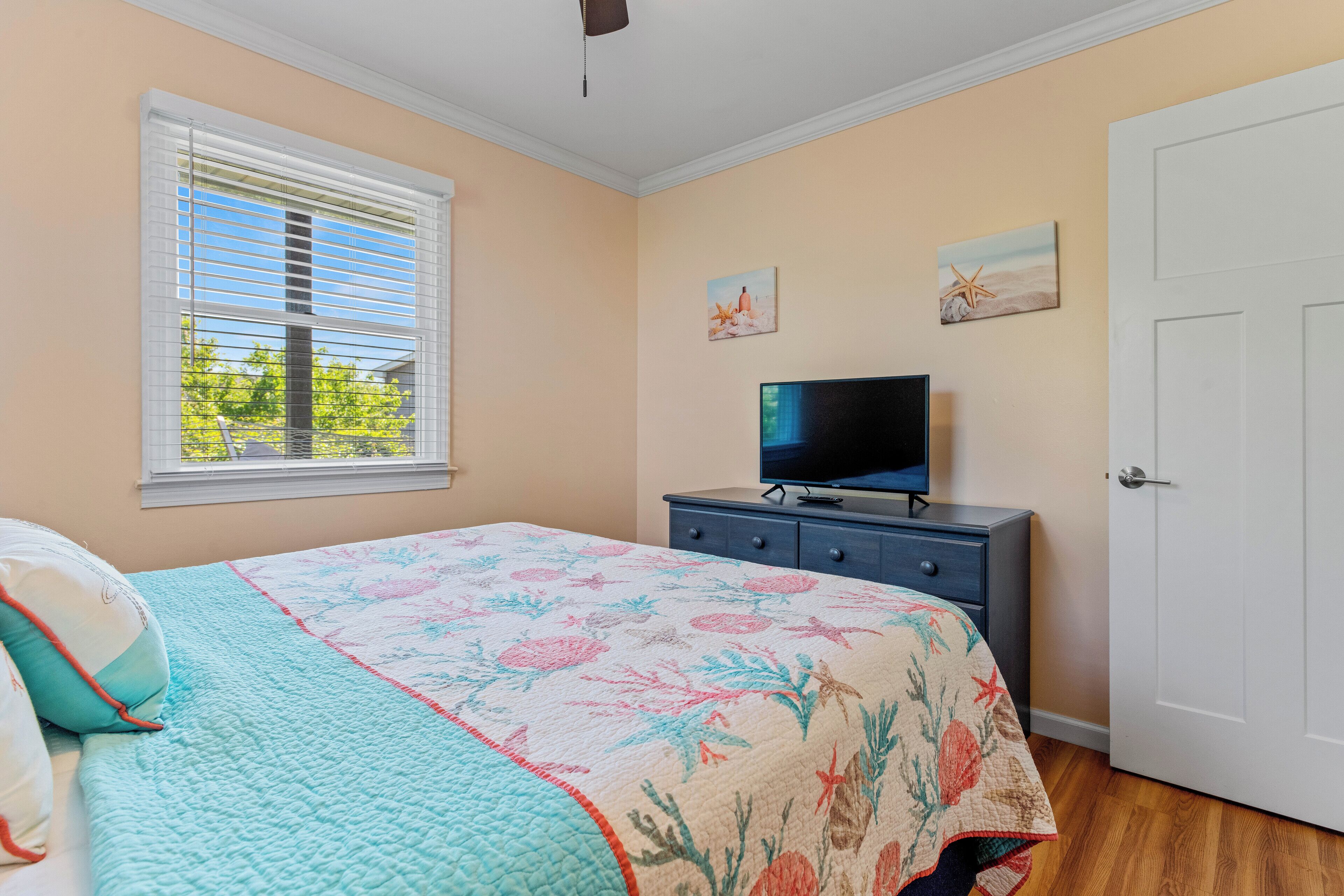 A bedroom with a bed, a TV on a dresser, a window with blinds, and a door.