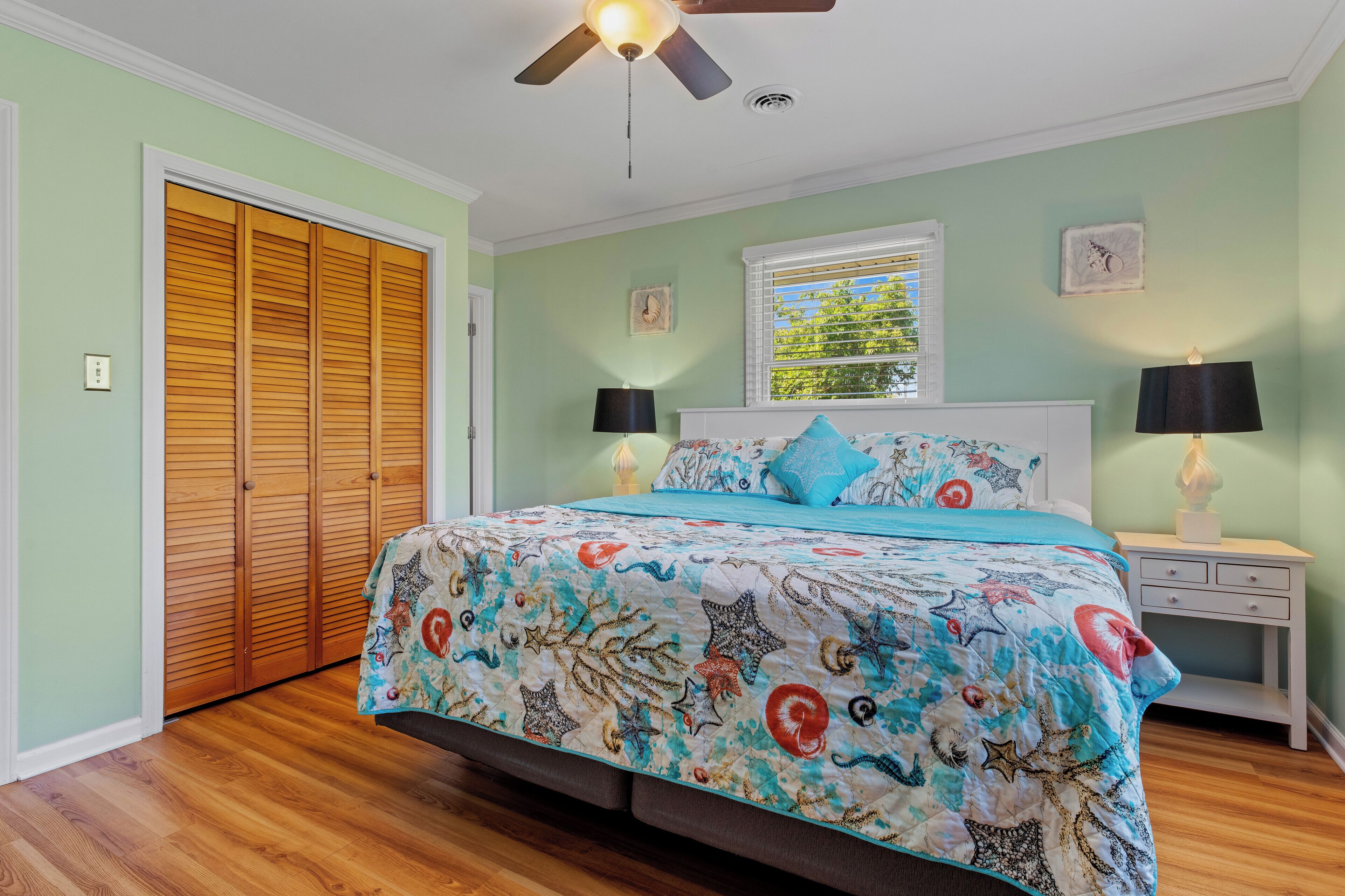 A bedroom with a bed, wooden closet, ceiling fan, nightstands, and a window with blinds.