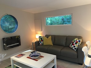 Uma sala de estar com sofá, mesa de centro, televisão, luminária e janela com vista para árvores.