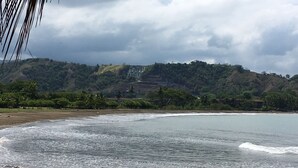 A beach with waves, a forested hill, and a cloudy sky. - Luxury Oceanfront Los Suenos Condo overlooking Herradura Bay (Los Suenos)