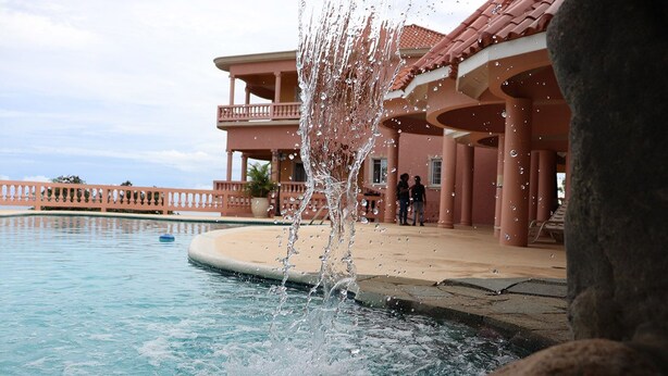 Una piscina con un elemento decorativo acquatico in primo piano, un edificio con un balcone sullo sfondo e un cielo sereno.