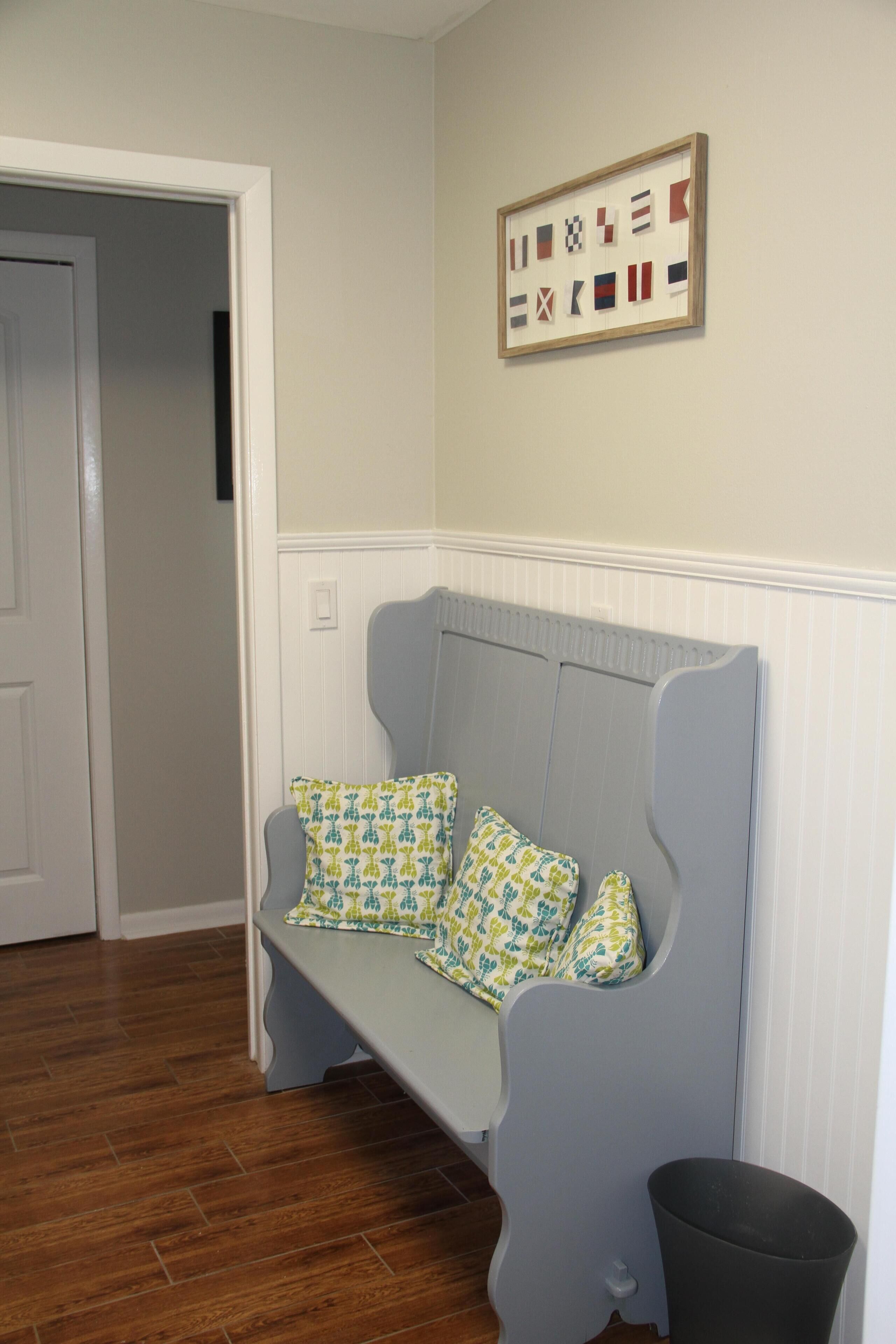 A room with a grey bench, patterned cushions, a wooden door, and a framed picture on the wall.