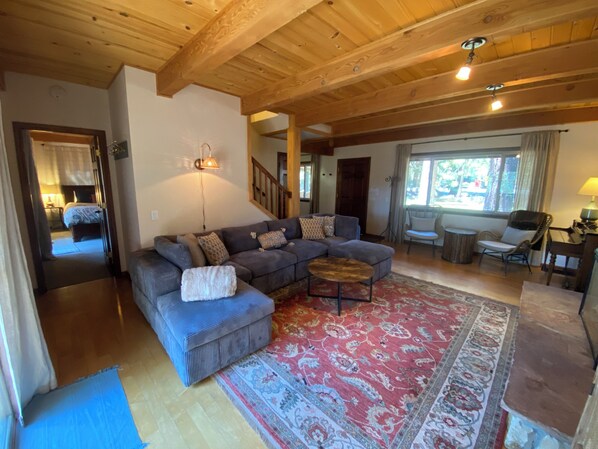 A spacious living room with a large sectional sofa, a coffee table, and a patterned rug. There is a staircase leading to another level, and a window with curtains.