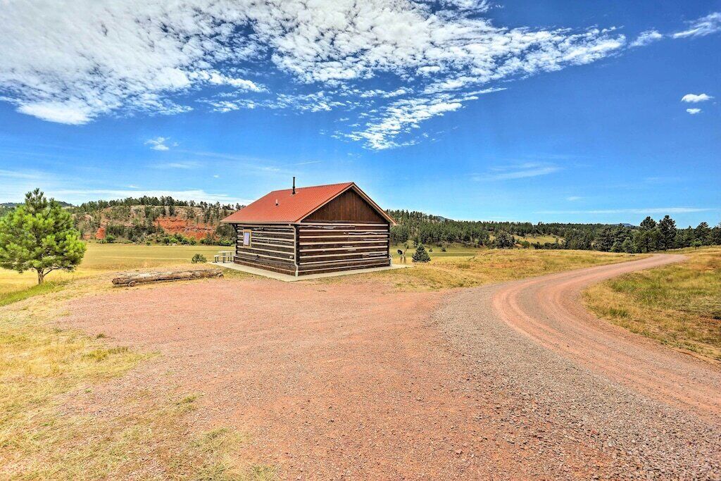 A wooden cabin with a red roof, surrounded by a gravel driveway and a dirt path, set in a rural landscape with trees and hills in the background.
