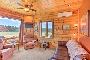 L’intérieur chaleureux d’un chalet avec des murs en bois, un ventilateur de plafond, une cheminée et une grande fenêtre offrant une vue panoramique.