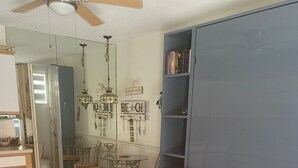 A living room with a beige sofa, a dining table with chairs, a bookshelf, and a ceiling fan.