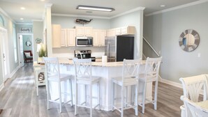 Una cocina moderna con una isla central, taburetes blancos tipo bar y armarios de madera clara.