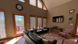 A spacious living room with a large black leather sofa, a wooden coffee table, and a television mounted on the wall. There are large windows with blinds, a red deck, and a view of a lake. - Heron Landing:  Escape to Lake Mayo.
Private cove, where nature abounds! 



 (Roxboro)