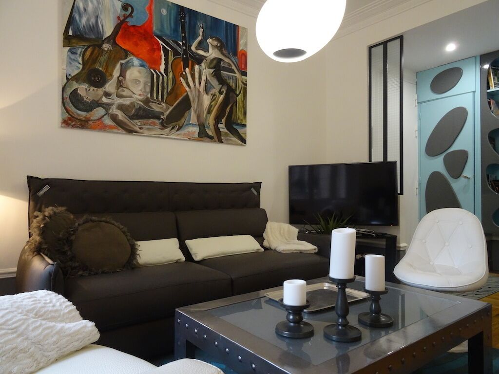 A modern living room with a dark sofa, a flat-screen TV, a glass coffee table with candles, and a painting on the wall.