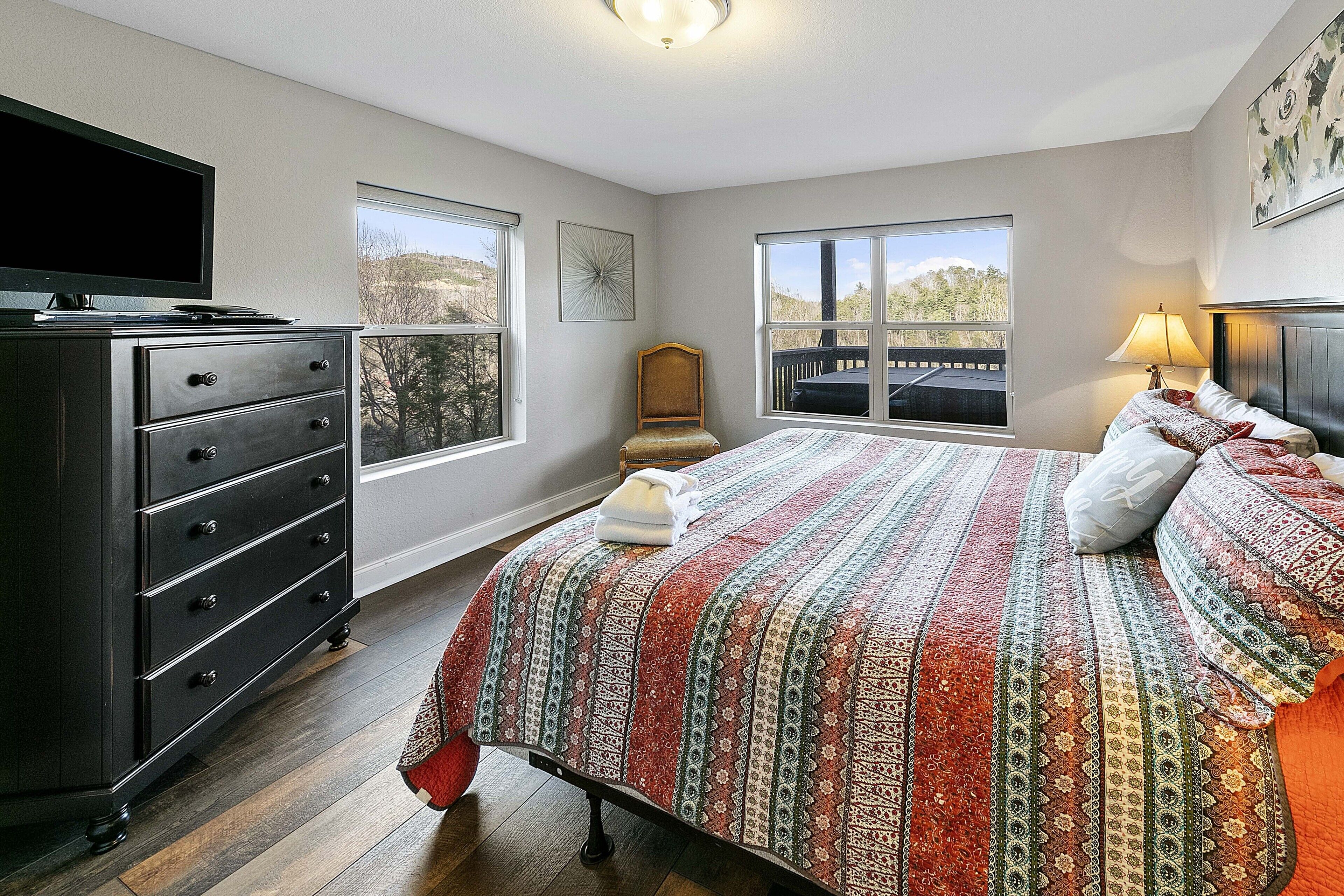 A bedroom with a bed, dresser, TV, windows, and a view of the outdoors.