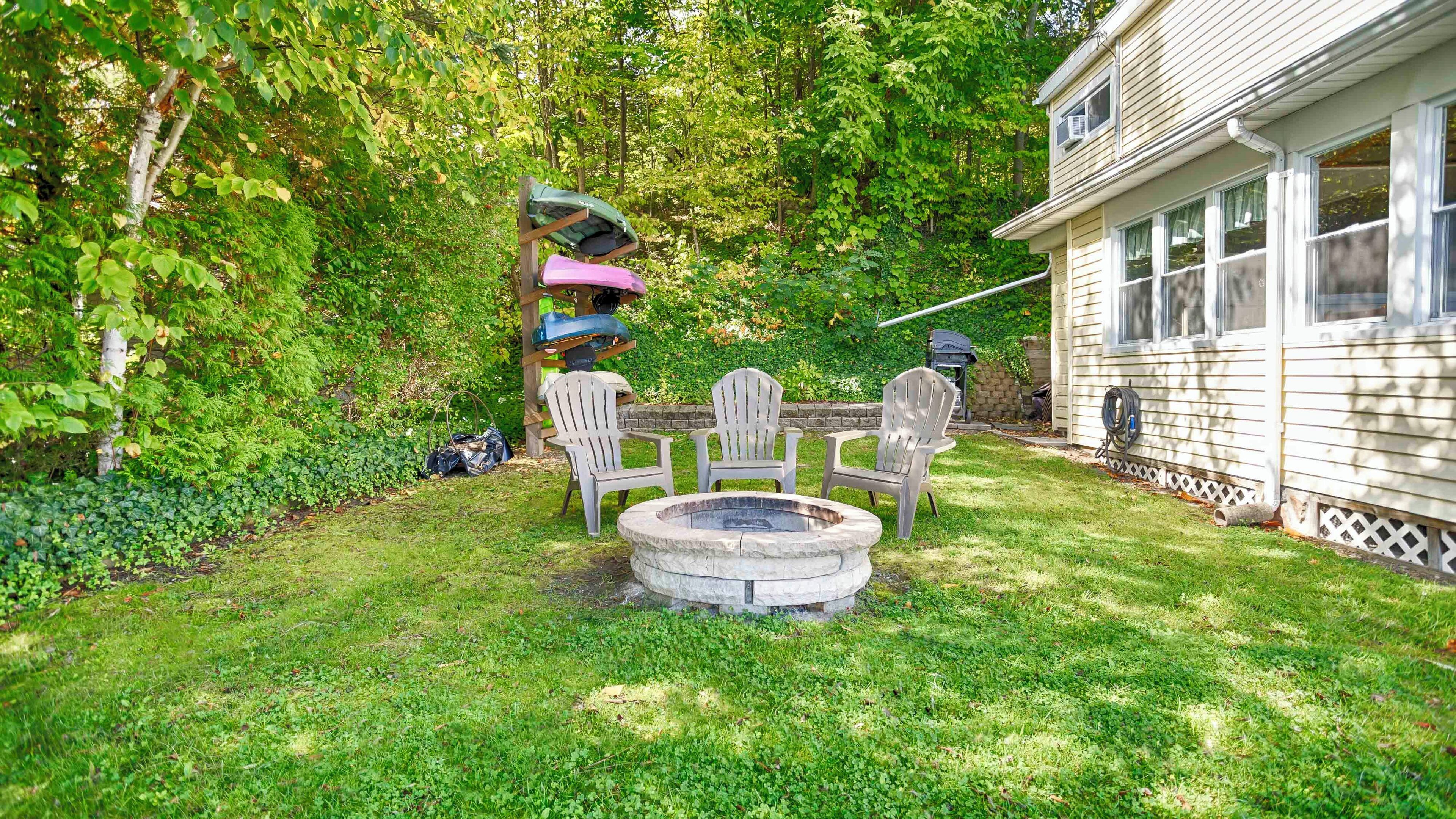 A backyard with a stone fire pit, three Adirondack chairs, and a wooden pergola with a green umbrella.