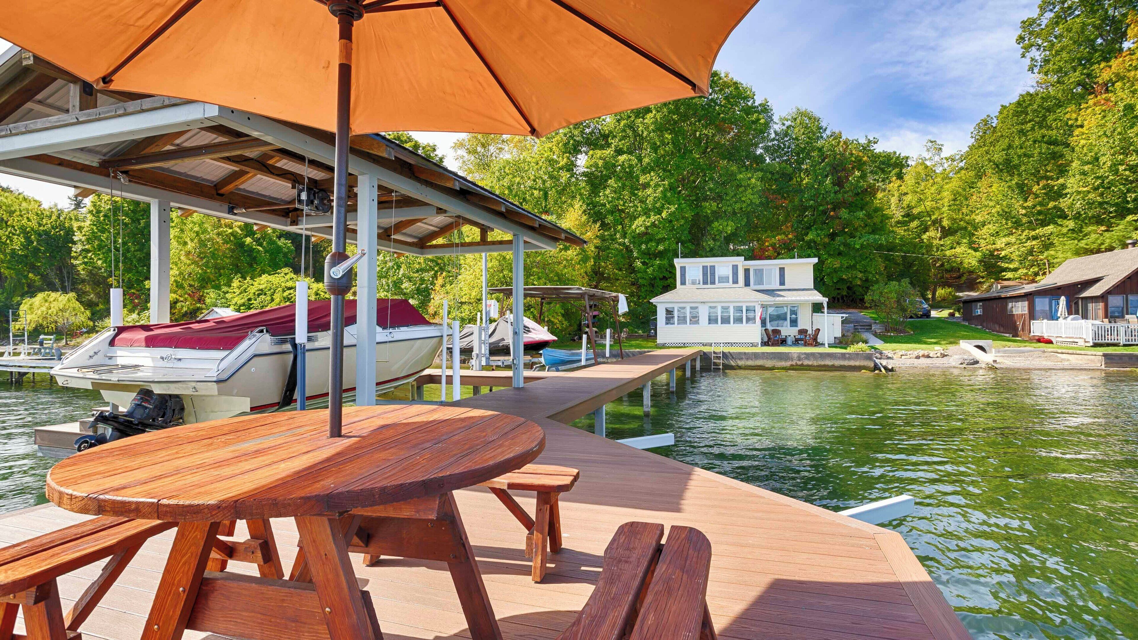 A wooden dock with a table and chairs, a covered area with an umbrella, and boats docked in the background.