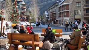 A snowy outdoor area with wooden benches, a Christmas tree, and buildings with balconies.