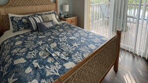 A bedroom with a large bed, a ceiling fan, a nightstand with a lamp, a wall-mounted fan, and a view of a balcony with a railing.