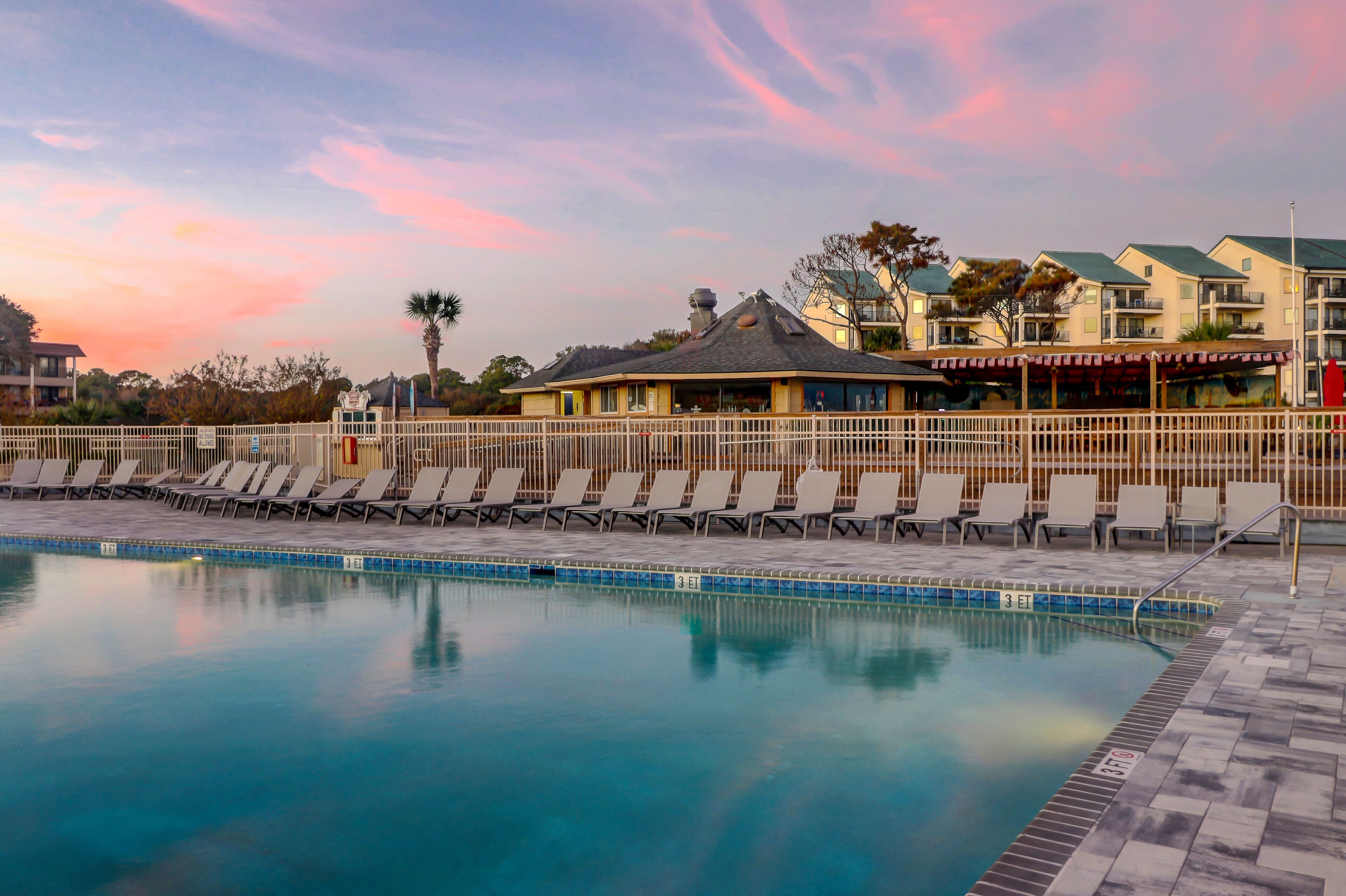 Une piscine avec des chaises longues, un bâtiment doté d’un toit végétalisé et un ciel au coucher du soleil.