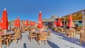 A deck with wooden tables and chairs, red umbrellas, and a building in the background.