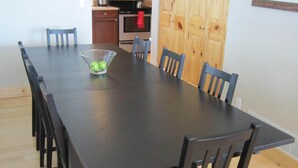 A dining room with a long table, chairs, a chandelier, a microwave, and wooden cabinets.