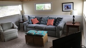 Een gezellige woonkamer met een grijze bank, een grijze fauteuil, een rieten pouf en een televisie. Er zijn twee bijzettafels met lampen, een raam met zonwering en een ingelijst schilderij aan de muur.