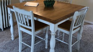 Een eettafel met witte stoelen, een groene kom en een lamp in een kamer met vloerbedekking en een radiator.