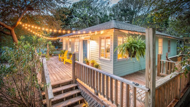Una terraza de madera con una escalera que lleva a una casa pequeña con porche, rodeada de follaje y luces cálidas.